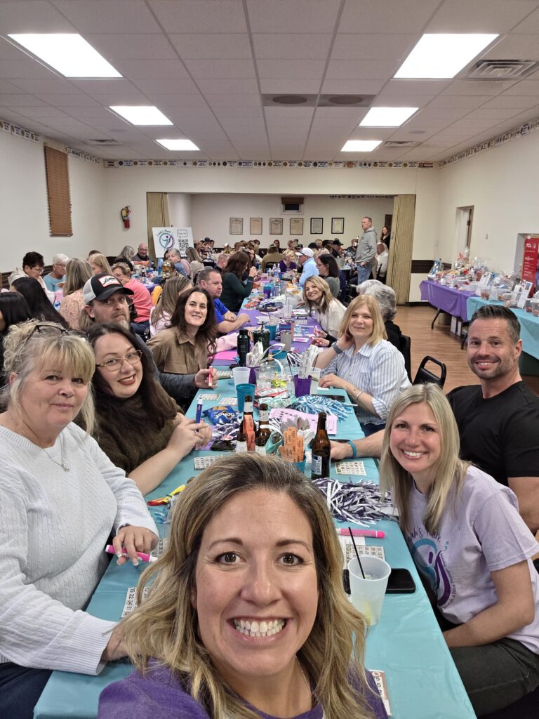 Selfie of numerous bingo players at tables with gift baskets in the background