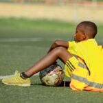 A young soccer player in a yellow jersey takes a break on the grass field with a soccer ball.