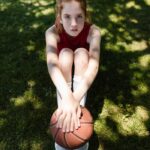 A young girl sits on grass holding a basketball, showing determination and focus in a summer setting.